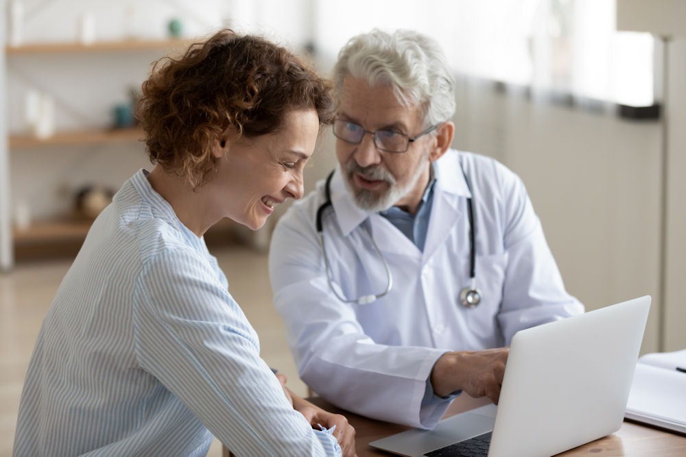 Doctor with stethoscope explains image on laptop screen to smiling woman.